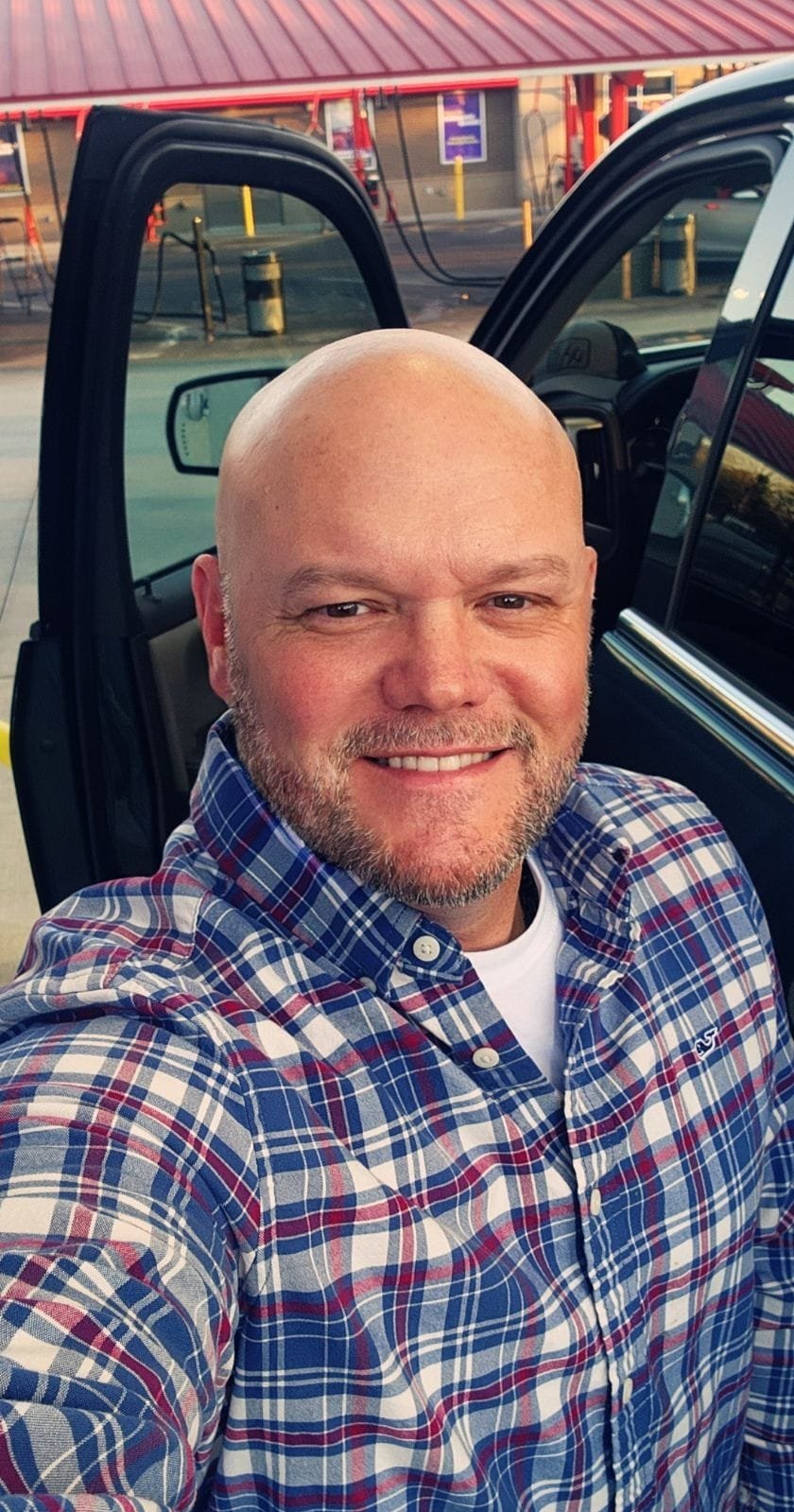 Bald man with reddish beard smiling at camera, wearing blue and red plaid shirt, sitting in truck cab with open door