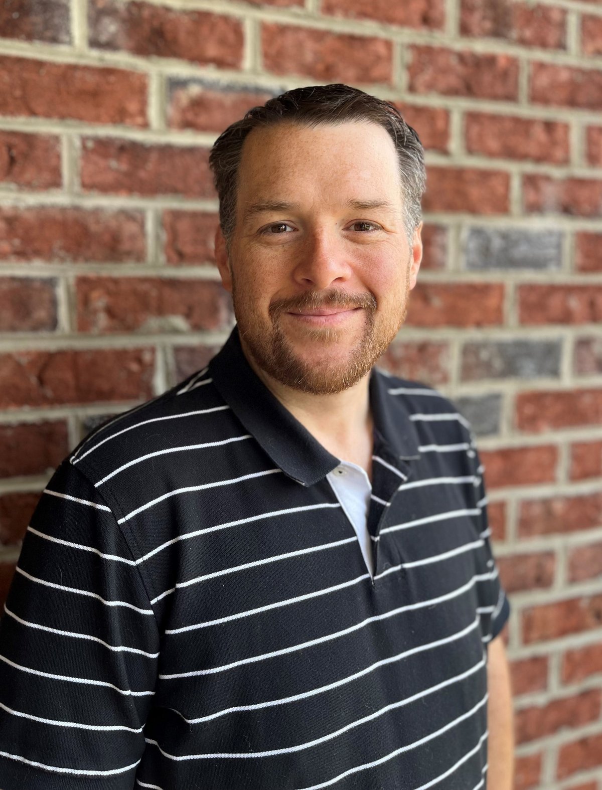 Man with dark hair and beard wearing navy striped polo shirt standing in front of red brick wall
